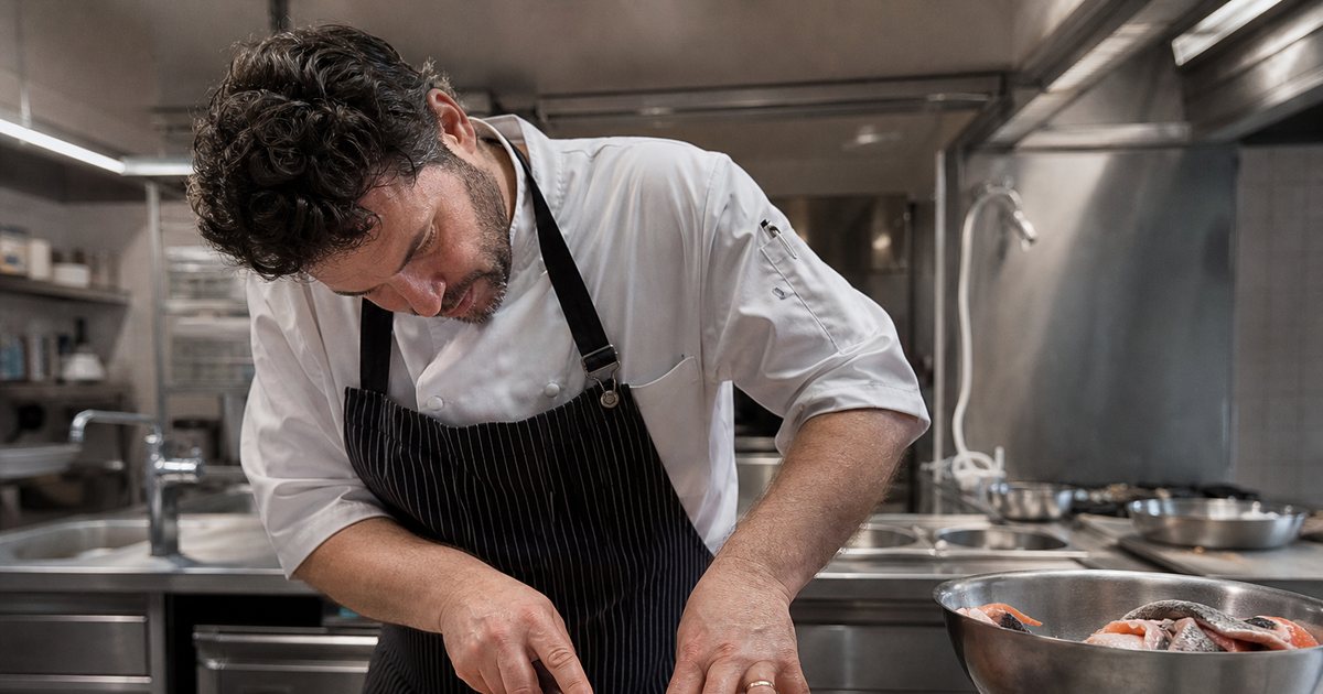 A chef in a stainless steel restaurant kitchen breaking down a whole salmon on a cutting board, trimmed skin and bones in a metal bowl beside a digital kitchen scale, overhead natural light